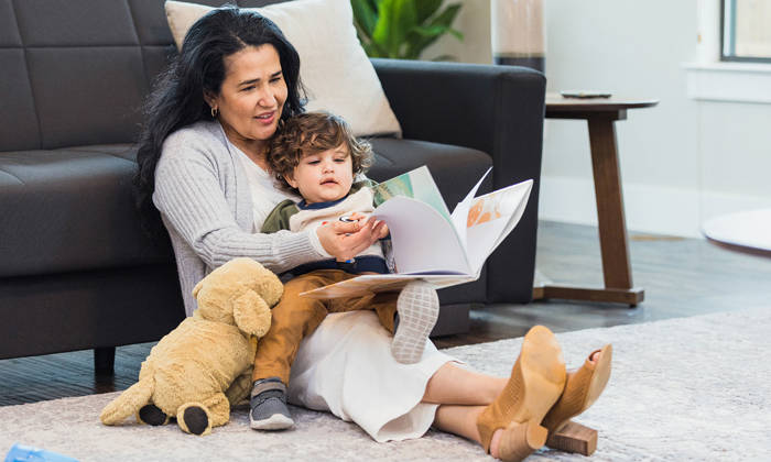 A grandmother and her toddler grandson sit on the floor of the living room and lean against the sofa for story time